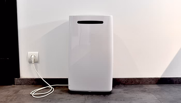 Un purificador de aire blanco está de pie, enchufado a una toma de corriente. Se encuentra en una habitación con suelo de baldosas grises y una pared blanca de fondo.