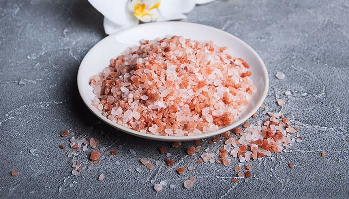 Un plato contiene sal rosada en grano grueso. Algunos granos están esparcidos sobre una superficie gris texturizada. En el fondo, hay una flor blanca decorativa.