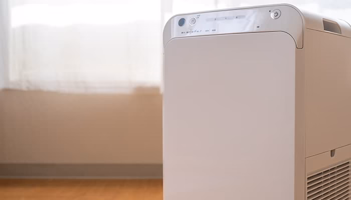Un purificador de aire está encendido en una habitación iluminada por la luz natural que entra por una ventana con cortinas blancas.