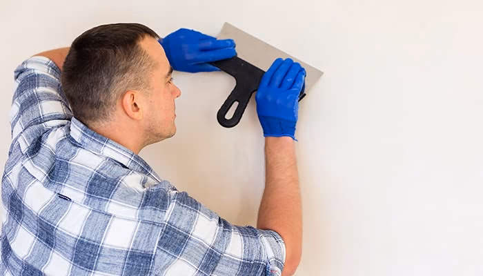 Hombre con camisa a cuadros, empuñando una llana, aplica yeso en una pared blanca dentro de una habitación. Lleva guantes azules de protección.