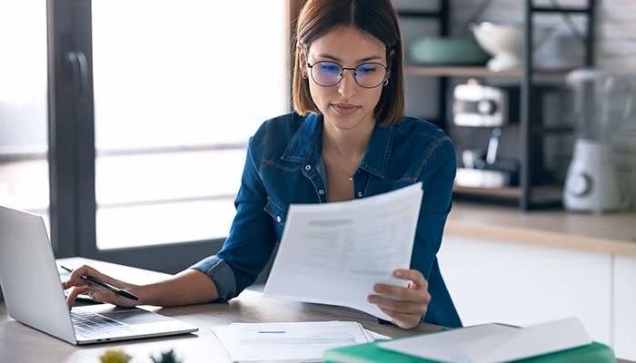 Una mujer sostiene documentos, revisándolos atentamente; está sentada frente a una computadora portátil en una cocina iluminada con estantes y utensilios al fondo.