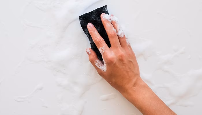 Una mano limpia una superficie blanca usando una esponja negra, esparciendo espuma de jabón en un entorno de limpieza doméstica.