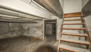 Unas escaleras de madera suben desde un oscuro sótano con paredes de cemento desgastadas. Se destacan tuberías metálicas en el techo y un suelo de concreto sin pulir.