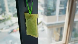 Un saquito verde cuelga de un gancho, colgando dentro de un edificio. Al fondo, se observa un entorno urbano con ventanas y edificios borrosos.