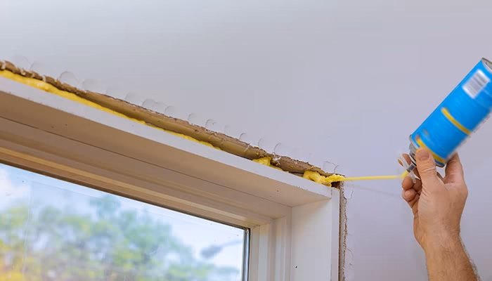 Una mano aplica espuma expansiva con aerosol alrededor de una ventana. La pared es blanca, y se observa el exterior a través del vidrio.
