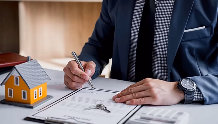 Un hombre escribe en un documento sobre una mesa con una pequeña casa de juguete al lado. Lleva traje y reloj, y hay calculadora y carpetas alrededor.