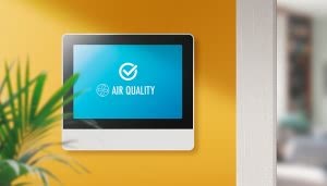 Pantalla mostrando "AIR QUALITY" e icono de ventilador, indicando buena calidad de aire. Está montada en una pared amarilla, con una planta verde cerca, en un entorno hogareño.