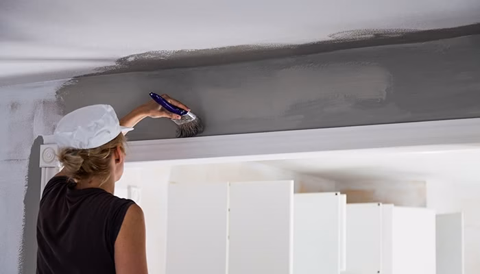 Una persona aplica pintura gris en el techo con una brocha. Lleva un gorro blanco. Al fondo, se observan gabinetes de cocina blancos.