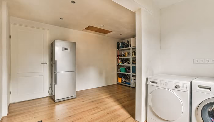 Un refrigerador metálico está en una habitación con piso de madera, junto a estanterías llenas de objetos. A la derecha, hay una lavadora y secadora.