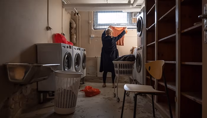 Una persona tiende ropa junto a una ventana en un cuarto de lavandería. La habitación cuenta con lavadoras, un lavamanos, cestos de ropa y estanterías de madera.