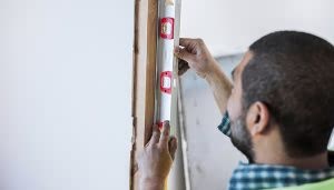 Un nivel de burbuja siendo ajustado por una persona en una pared. La persona lleva una camisa de cuadros y el entorno es un área de construcción o renovación.