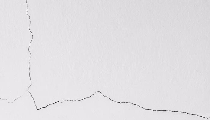 Una pared blanca muestra una delgada grieta negra horizontal, recorriendo y fragmentando la superficie, rodeada por un fondo liso y minimalista. No hay texto presente en la imagen.