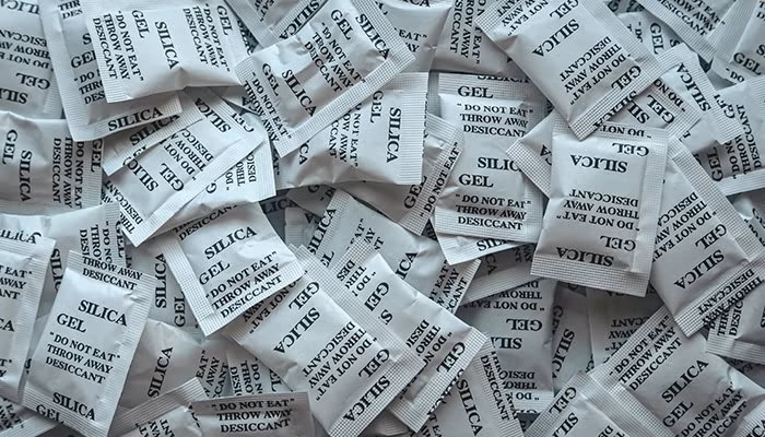 Paquetes de gel de sílice (pequeñas bolas antihúmedad) se encuentran esparcidos, con inscripciones en inglés: "Silica Gel," "Do Not Eat," "Throw Away," "Dessiccant," en un entorno de luz tenue.