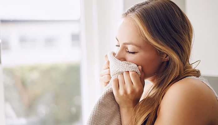 Una persona sostiene y huele una manta suave, disfrutando de su aroma en un espacio bien iluminado con una ventana y vegetación visible al fondo.