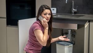 Una persona hablando por teléfono parece preocupada. Está frente a un fregadero con la puerta del gabinete abierta. El contexto es una cocina moderna.