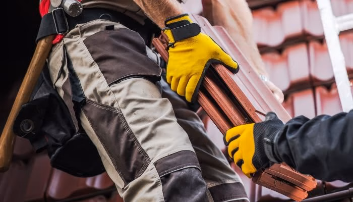Un trabajador con guantes amarillos sostiene tejas mientras otro le ayuda; están sobre un techo con tejas rojas y una escalera está cerca.