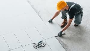 Trabajador colocando baldosas grises en el suelo, usando guantes y casco amarillo. Está en un espacio interior en construcción, sobre una superficie de cemento.