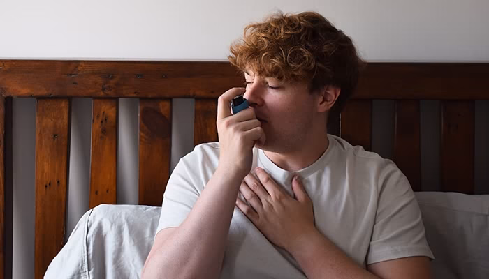 Un hombre con camiseta blanca usa un inhalador, llevando una mano al pecho, mientras está sentado en una cama con cabecera de madera oscura.