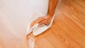 Una mano pasa un paño blanco sobre un piso de madera, limpiando un derrame líquido. El entorno es un ambiente interior con suelo de madera clara.
