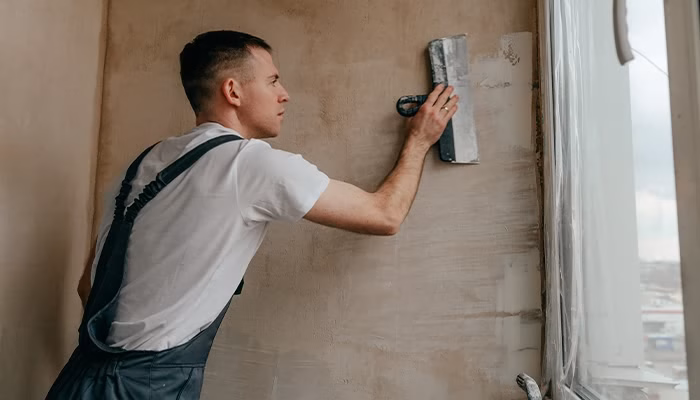 Un hombre alisa una pared con una espátula grande. Está concentrado en su trabajo, usando un mono gris y camiseta blanca, en una habitación iluminada por luz natural de una ventana.