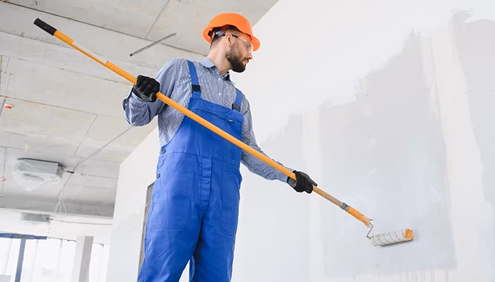 Un trabajador sostiene un rodillo de pintura y pinta una pared. Lleva un casco naranja y guantes negros dentro de un edificio en construcción.