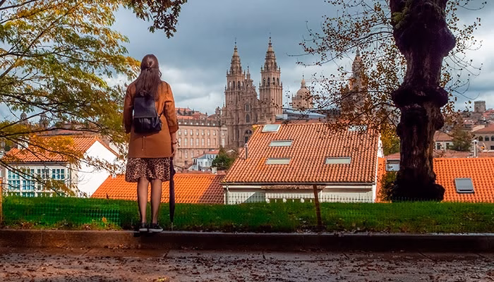 Una persona de pie observa la catedral desde un parque, rodeada de árboles y edificios con techos rojos. El cielo está nublado. Lleva una mochila y un paraguas.