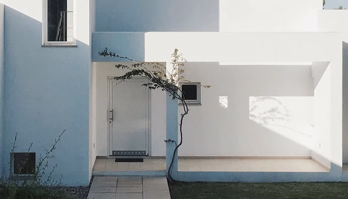 Una fachada moderna de color blanco y azul. Una puerta y una pequeña ventana están centradas. Una planta delgada crece junto a la entrada, proyectando sombras leves.