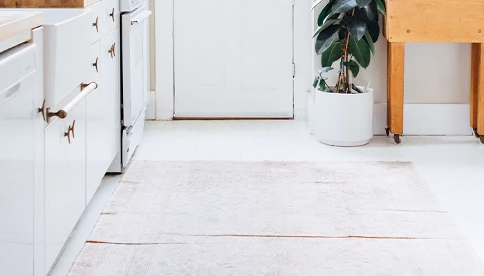 Una alfombra beige se extiende en el suelo de una cocina blanca, con gabinetes a la izquierda y una planta en maceta junto a una puerta al fondo.