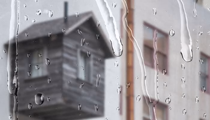 Gotas de lluvia resbalan por un vidrio mientras, al fondo, se observa un edificio de madera y otro de ladrillo difuminados. El contexto es un día lluvioso.