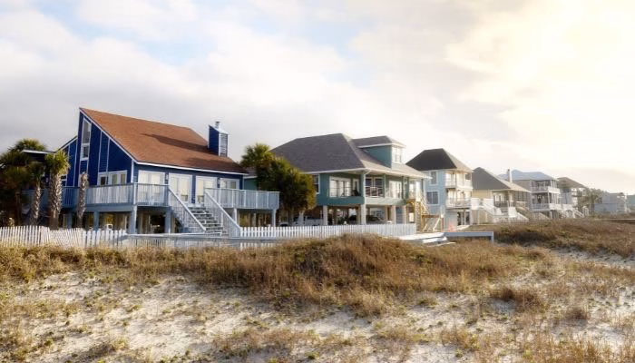 Casas de playa elevadas permanecen tranquilas en un ambiente costero, rodeadas de dunas de arena y vegetación escasa, con un cielo parcialmente nublado y luz suave de atardecer.