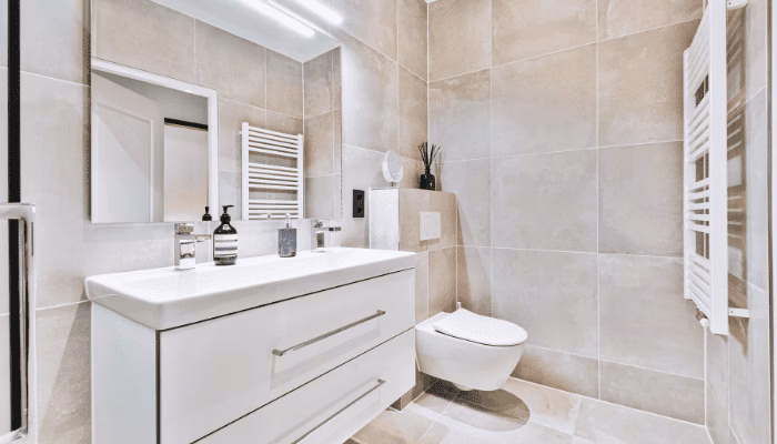 Un lavabo moderno con grifos y dispensadores de jabón se encuentra en un baño revestido de azulejos beige. A su lado, un inodoro suspendido y un espejo iluminado.