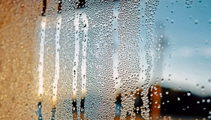 Pequeñas gotas de agua resbalan por una ventana de vidrio, formando patrones irregulares; al fondo, un paisaje borroso con un cielo azul y edificios difuminados.