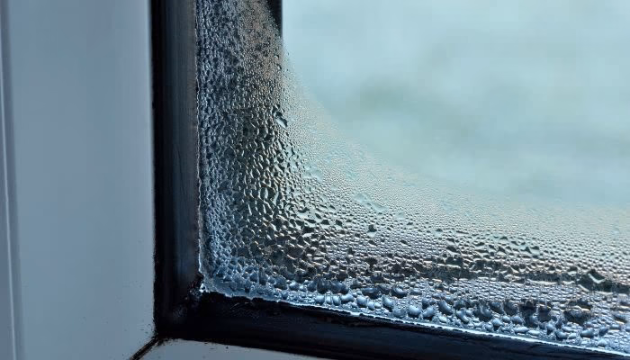 El cristal de una ventana muestra condensación y gotas de agua, acumulándose por la humedad en la esquina inferior. Alrededor, un marco de ventana blanco en un día frío.