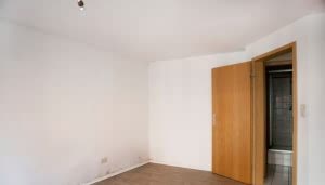 Una habitación vacía tiene una puerta de madera abierta que conduce a un baño. Las paredes son blancas y el suelo es de madera. Hay humedad visible en una esquina.