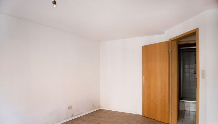 Una habitación vacía tiene una puerta de madera abierta que conduce a un baño. Las paredes son blancas y el suelo es de madera. Hay humedad visible en una esquina.