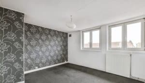 Una habitación vacía con alfombra gris y pared decorada con papel tapiz floral en el lado izquierdo. Tres ventanas grandes iluminan el espacio desde la derecha; un radiador está debajo.