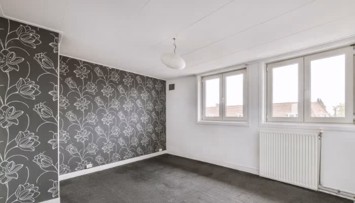 Una habitación vacía con alfombra gris y pared decorada con papel tapiz floral en el lado izquierdo. Tres ventanas grandes iluminan el espacio desde la derecha; un radiador está debajo.