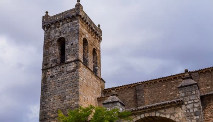 Una torre de piedra se alza, con ventanas arqueadas, en el contexto de un cielo nublado. Alrededor, vegetación verde y un arco de entrada complementan la estructura.
