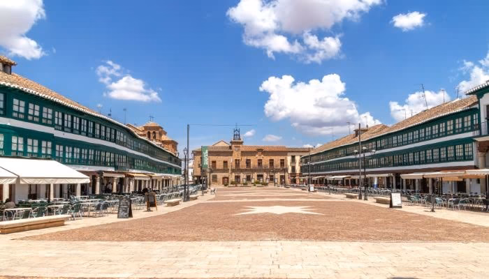 Imagen de la plaza de Ciudad Real con edificios de fachadas verdes a ambos lados, mesas al aire libre y cielo azul con nubes blancas. Al fondo, un edificio histórico destaca en el centro.