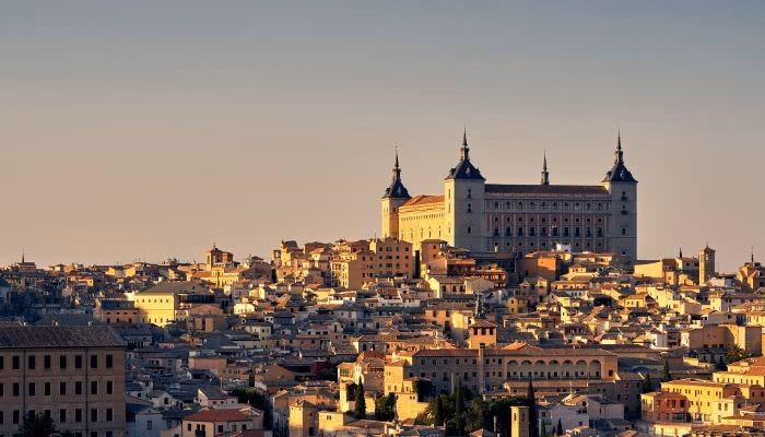 Un gran edificio histórico con torres se eleva sobre una ciudad antigua con numerosas casas en tonos cálidos. El cielo está despejado, sugiriendo un día soleado.