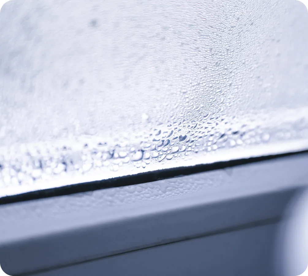 Gotas de agua se acumulan en una ventana empañada, creando un patrón de condensación. La luz difusa ilumina el vidrio, sugiriendo un ambiente frío y húmedo.