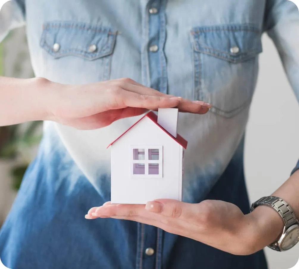 Una persona sostiene con ambas manos una pequeña maqueta de casa blanca con techo rojo, colocándola en el centro de su pecho. Lleva una camisa de tela vaquera. Gas radón.