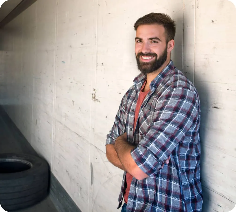 Un hombre con camisa a cuadros cruzado de brazos, sonríe mientras se apoya en una pared clara. A su lado hay una llanta en un espacio cerrado.