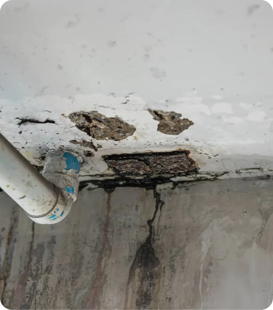 Una tubería de plástico blanca con pintura azul desgastada sobresale, junto a un techo deteriorado con humedad visible. Contexto: paredes grises con manchas de agua y grietas.