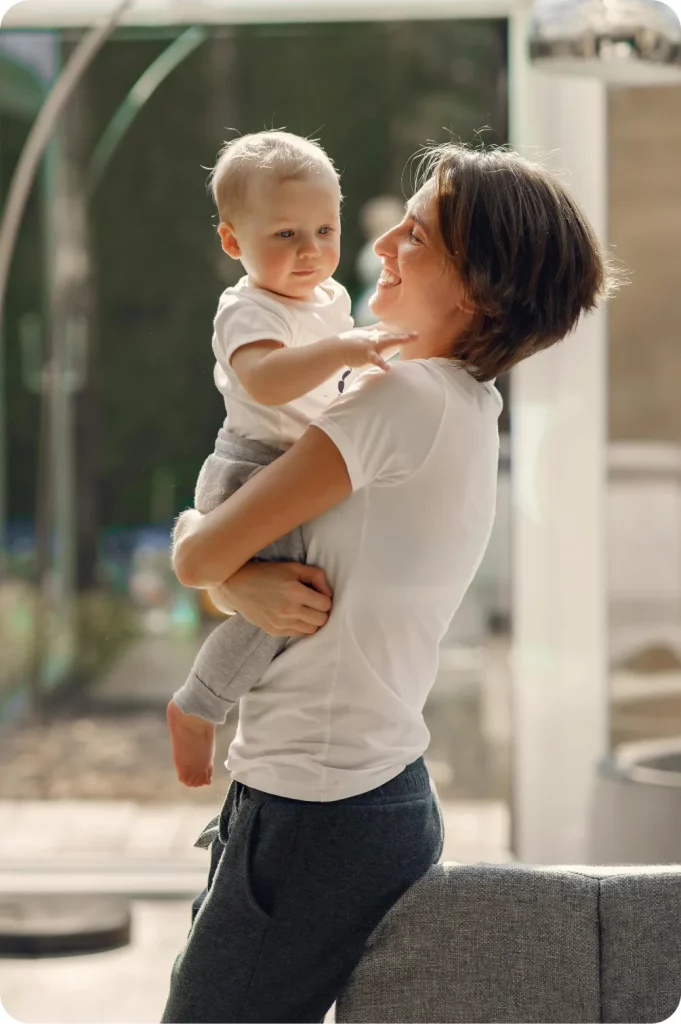 Sanysec - Tratamientos definitivos contra la humedad en Pontevedra Una persona sostiene a un bebé mientras sonríen mutuamente, en un entorno iluminado por la luz natural, con un fondo de ventanales y muebles modernos.