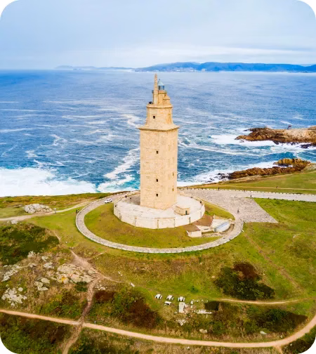 La torre se alza imponente, rodeada de un sendero circular. El océano Atlántico se extiende detrás, con olas chocando contra las rocas cercanas. El cielo está despejado.