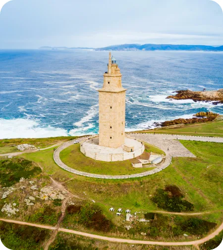La torre se alza imponente, rodeada de un sendero circular. El océano Atlántico se extiende detrás, con olas chocando contra las rocas cercanas. El cielo está despejado.
