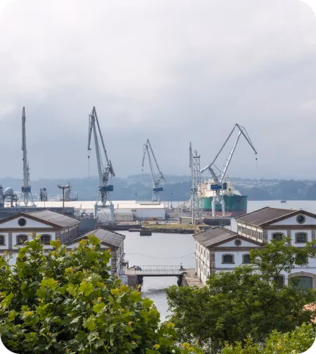 Grúas portuarias se elevan inactivas en un astillero, rodeadas de edificios industriales y vegetación. El cielo está nublado y el agua del puerto refleja el entorno.